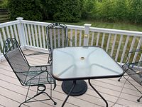 Photo showing square patio table with frosted glass top and two metal mesh chairs with decorative backs placed on deck
