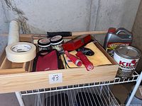 Wooden tray with paint roller handles, masking tape, small paint cans, red toolbox, microfiber towels, and gloves arranged on a white wire shelf.