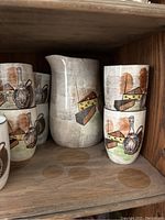 Photo showing the pitcher and stack of drinking glasses with cheese and wine bottle painted design on a wooden shelf.