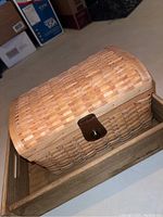 Light-colored woven storage basket with a brown latch, sitting inside a rectangular wooden tray with side handles on a beige floor.