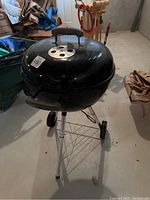 Closed black Weber charcoal kettle grill with vented lid and handle, standing on chrome legs with wheels and wire rack.