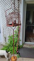 Full view of the metal birdcage mounted on the tall stand placed outdoors on a patio with greenery in the background.
