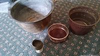 Four metal items arranged on carpet: large brass planter with floral engravings, smaller copper planter with beaded rim, copper-plated candle ring, small brass goblet.