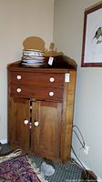 Vintage wooden corner cabinet with two upper drawers and a lower cupboard with double doors, ceramic knobs visible. Scratches and signs of use on wood surface. Plates and bowls shown on top.
