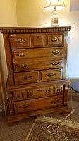Wooden 4 drawer dresser shown from front angle with metal handles and decorative molding at the top, sitting on carpeted floor with lamp on top.