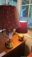 Two brass lamps on wooden surface near window, both with burgundy fabric lampshades with swirling patterns.