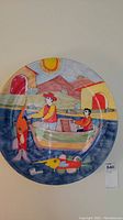 Front view of the round ceramic plate showing colorful fishing scene with two figures in a boat, fish, water, sun, and buildings in background.