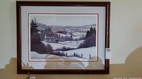 Framed print of winter countryside showing snow-covered landscape, houses, trees, and hills in muted colors.