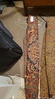 Rolled-up rug leaning against suitcase and furniture, showing the detailed pattern and label on top.