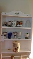 Photo of white wooden wall shelf holding ceramic egg coddlers, a small canister with lid, a glass jar with lid, and a ceramic container with metal clasp at bottom shelf.