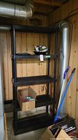 Tall black plastic shelving unit with five shelves placed against a wooden wall in a basement or storage area. Items on shelves include a box and miscellaneous household objects.