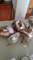 Overview photo of five silver plated items on a table, including a lidded serving dish, gravy boat, cocktail shaker, teapot, and bowl with tarnishing and patina.