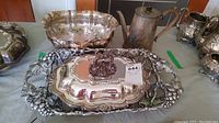 Silver-plated serving dish with lid featuring grapevine motif and ornate handle, next to tarnished silver-plated bowl and antique copper teapot with handle and spout.