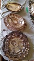 Image showing three silver-plated trays, one large round ornate tray holding three miniature ashtrays and two small serving dishes, and a gravy boat on a scalloped tray, all tarnished and needing cleaning.