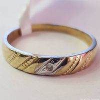 Close-up photo showing the ring's yellow gold band with diagonal textured patterns and a small diamond set in white gold accent area.