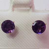 Close-up image of the front view of two round purple amethyst earrings set in 14K yellow gold prong mounts, displayed on a white textured jewelry holder.