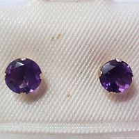 Close-up front view of the pair of round faceted purple amethyst stud earrings set in 14K yellow gold prongs on a cream fabric display
