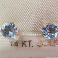 Close-up photo of pair of round aquamarine gemstones set in 14K yellow gold prong settings on a cream card with text '14 KT. GOLD'.