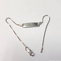 Silver ID bracelet laid out flat showing rectangular ID plate, slender box chain and lobster clasp closing mechanism.