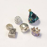 Six silver-tone Pandora style beads displayed on a white background with visible details including a Christmas tree bead with colored rhinestones, a round bead covered in clear rhinestones, a silver spherical clip bead, a lavender crown bead with gold details, and a silver book bead with a black cross.