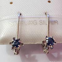 Front view showing two sterling silver earrings with blue sapphires in six-prong settings. Hook and clip fastening visible.