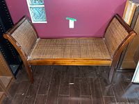 Full front view of vintage cane bench against burgundy background showing wooden frame and woven cane seat and arm panels.