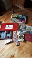 Photo of the various boxed board games and game components arranged on floor, including Stock Ticker, Hararddopoly, Masterpiece, plus cards and dice.