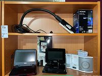 Two portable DVD players on shelf with Sony S-frame photo frame box behind, small white music box, flexible desk lamp, and boxed NOMA lamp.