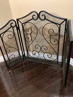 Full view of three-panel folding fireplace screen with iron scroll design and mesh backing standing on wood floor near beige wall.