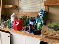Wide view of gardening care items on a wooden workbench including fertilizer bottles, red gas container, spray bottles, bug spray, and brown leaf bags stacked on the right