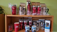Wide view of assorted new travel mugs, coffee mugs, and tumblers on wooden shelves including red and chrome travel mugs, red ceramic mugs, and blue logo mugs.