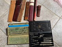 Top-down view of two cases with vintage metal drawing compasses, dividers, and instruments spread inside. Cases include green fabric-lined and black hard case with gold emblem.