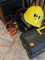 Photo showing yellow hard hat on top of a black plastic toolbox with orange power cable and an orange hacksaw visible