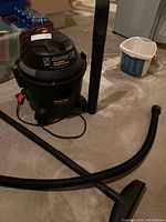 Shop-Vac 5 gallon wet/dry vacuum with hose and floor tool attachment on the floor, showing whole unit and accessories.