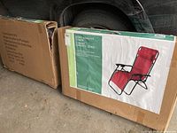 Photo of two unopened cardboard boxes containing Zero Gravity Chairs in front of a vehicle tire, one box shows product picture and highlights powder coated steel frame and textilene fabric.