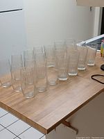 20 clear drinking glasses displayed on a wooden table in two groups of 10, showing two different designs: one set with subtle etched pattern, one set with vertical etched lines.