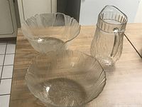 Photo of two clear crystal salad bowls and one clear crystal jug arranged on wooden table, showing floral and scalloped design on bowls and ribbed texture on jug.