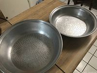 Two round metal strainers placed on a wooden table. Each strainer has multiple small holes on the base for draining liquids from vegetables and fruits.