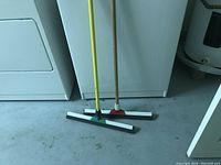 Photo shows two floor wipers standing side by side with long handles and rubber blades suitable for wiping messes on tile floors.
