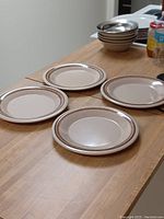 Four beige ceramic dinner plates with black and brown ring decorations placed on a wooden surface. Plates grouped closely to show they are part of a set.