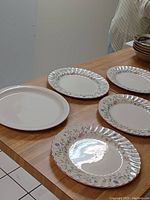 Five ceramic plates on wooden surface: four with floral scalloped rim designs, one plain white plate.