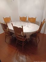 Six wooden dining chairs arranged around a table with a white patterned cover in a room with hardwood floors.