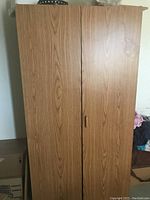 Photo of the front of a brown wooden closet cabinet with two doors and wooden handles. The cabinet is standing on the floor against a wall.