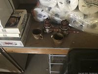 Photo of two small metal coffee boilers with wooden handles and decorative bands, accompanied by two small matching metal cups, taken on a shelf.