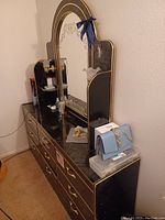 Black dresser with gold trim, mirror, and six drawers. Items on top include a blue purse and personal care products.