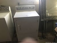 White electric clothes dryer with front door and top control panel, placed next to a washing machine and mirror frame.
