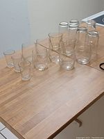 16 clear glass drinking glasses of assorted sizes and ribbed designs on a wooden table.
