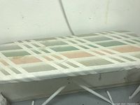Close-up of the ironing pressing table surface showing fabric cover with pastel stripes and white metal frame with adjustable legs.
