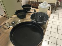 Photo shows electric juicer, aluminum bundt cake mold, large black round tray with glass lid, and two black plastic strainers with handles placed on a wooden table.