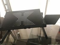 Photograph showing a black adjustable laptop table with a perforated 'X' pattern surface, foldable legs, and an additional side platform for extra space.